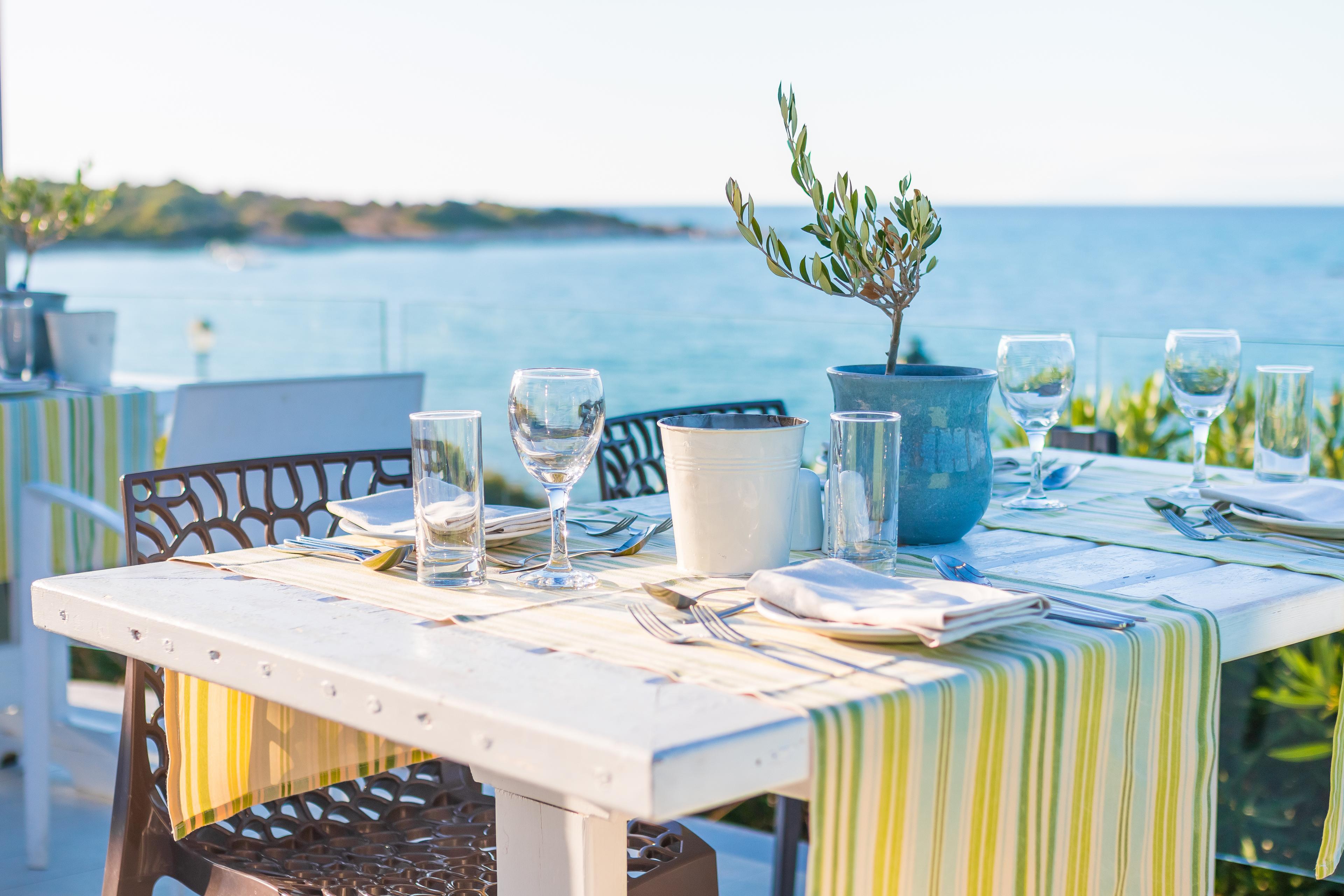 A Greek seaside restaurant table set for service, with an olive sapling centerpiece and the sea visible behind.