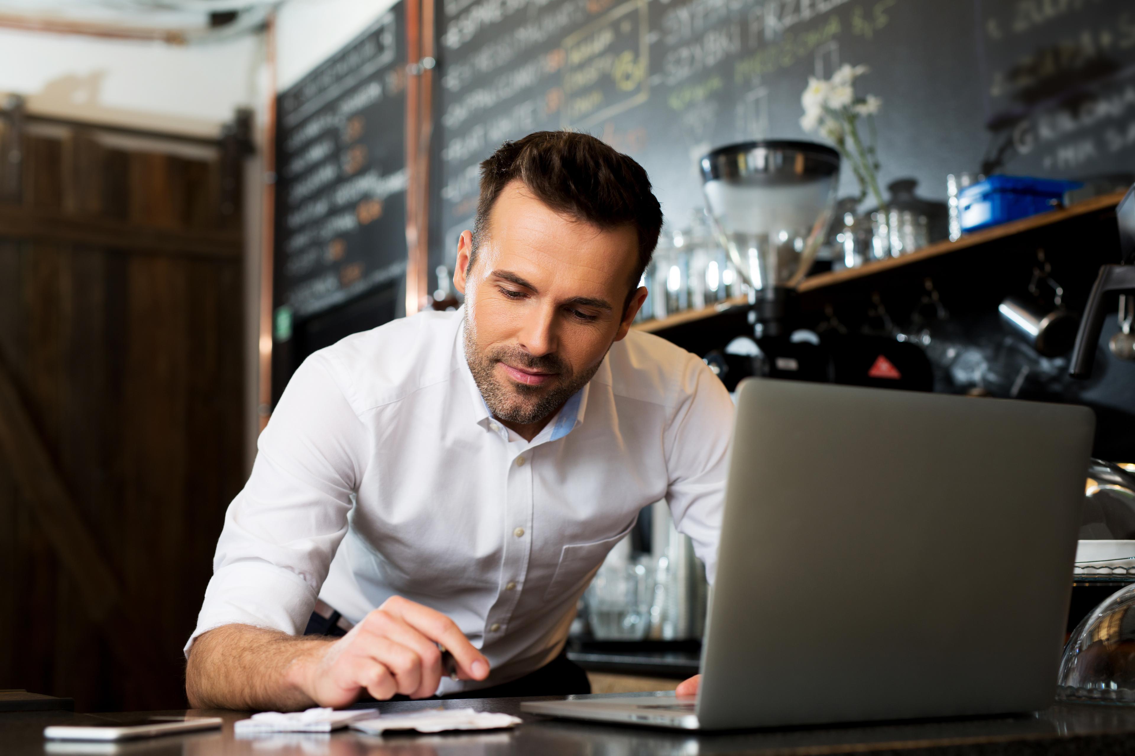 A cafe manager at the counter working on a laptop, with a chalkboard menu behind.